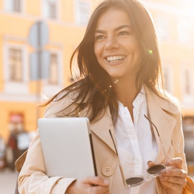 Happy brunette woman in jacket holding laptop computer and looking away outdoors